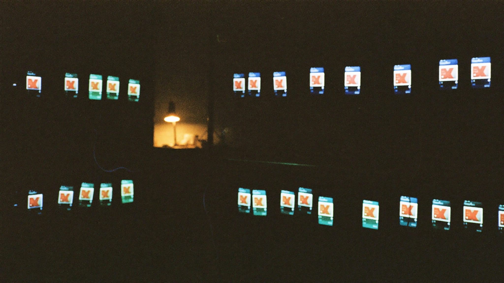Two long rows of phones glowing in a dark room, every screen showing the same red cross, with one amber lamp lit in a window between them