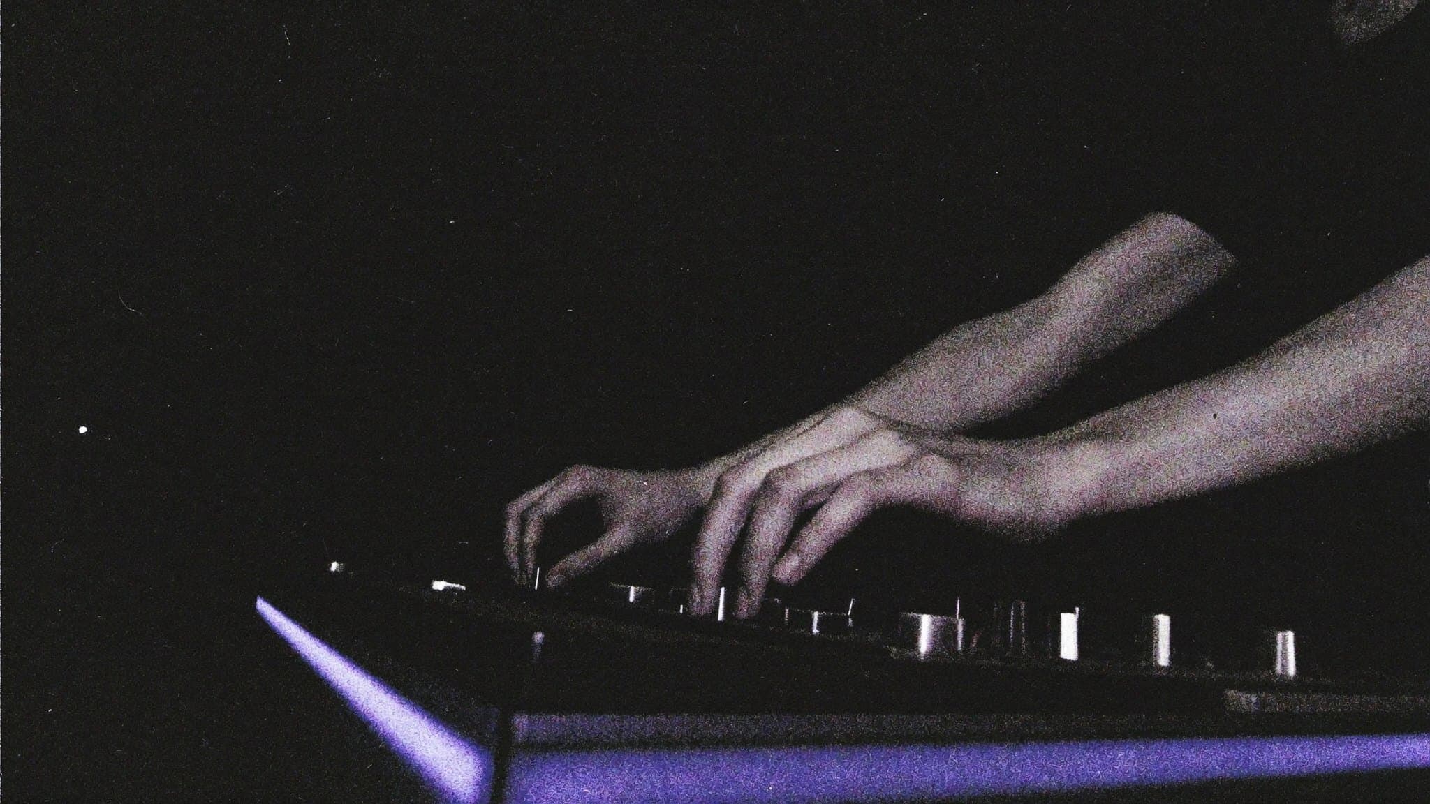 A producer's hands on a mixing console fader in near darkness, edged in violet light